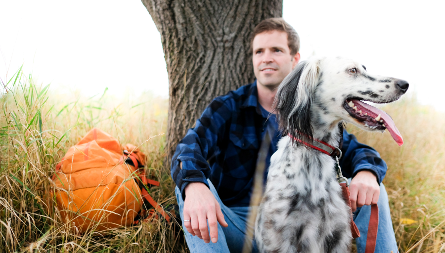 Mann sitzt mit orangenem Rucksack vor einem Baum auf der Wiese. Sein Hund sitzt vor ihm.
